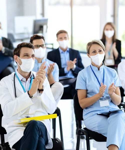 Large group of doctors and business people applauding while attending educational event during coronavirus pandemic. Focus is on male doctor.